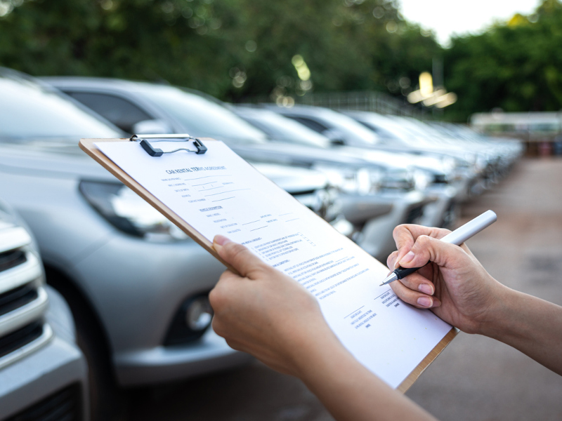 A woman writes on a clipboard while standing in front of a car lot filled with various vehicles.